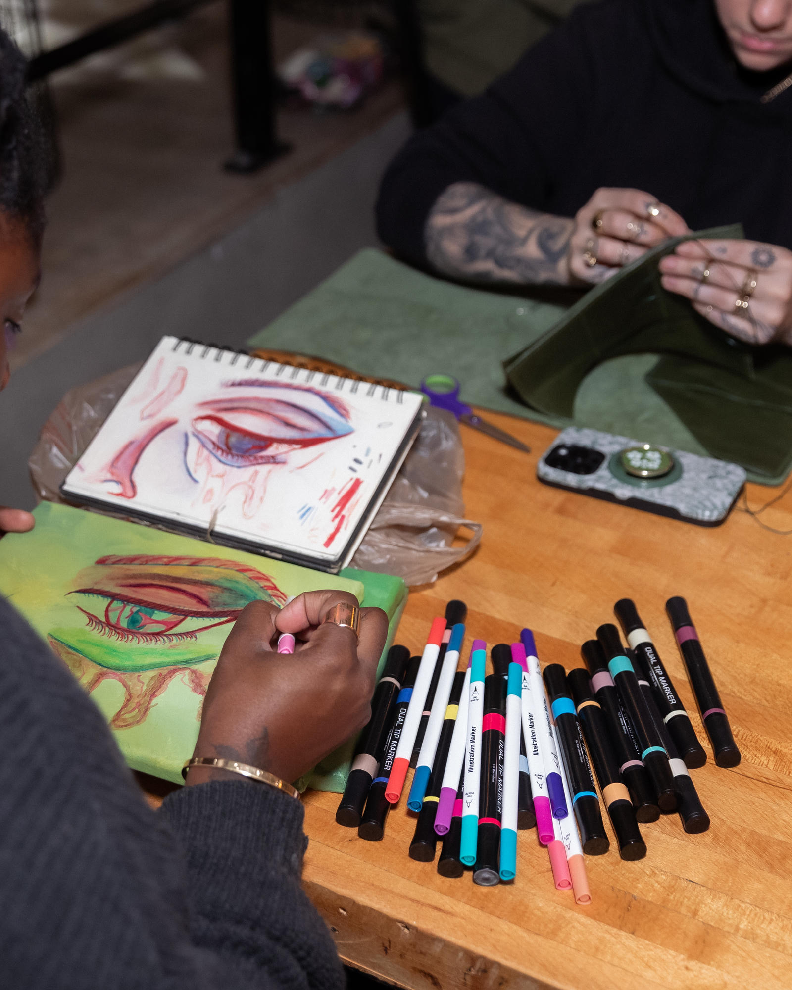 Colorful eye drawing on a green canvas beside its sketchbook study and markers